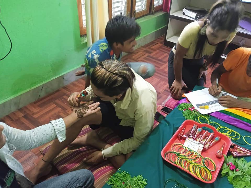 group of people gathered indoors, one is drawing a plant pattern on another's palm while others are looking on as one draws on paper