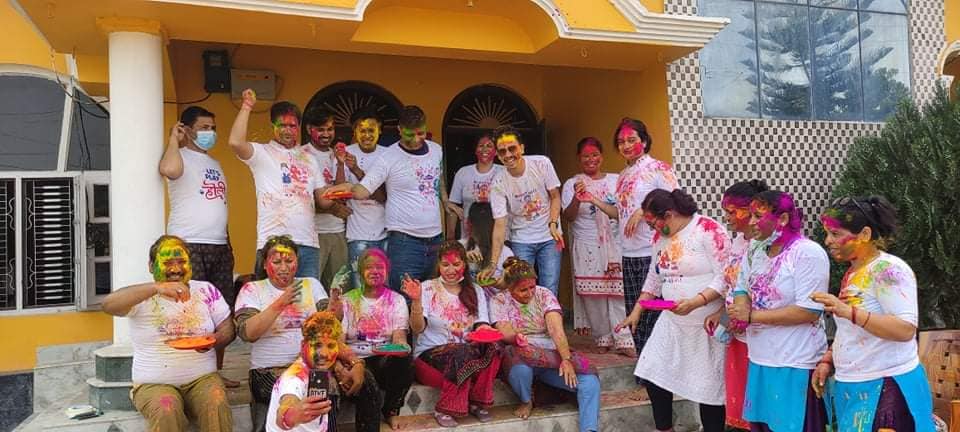 A group of people sitting and standing in front of an orange-colored house. All the people are wearing white tops and are covered in Holi colors