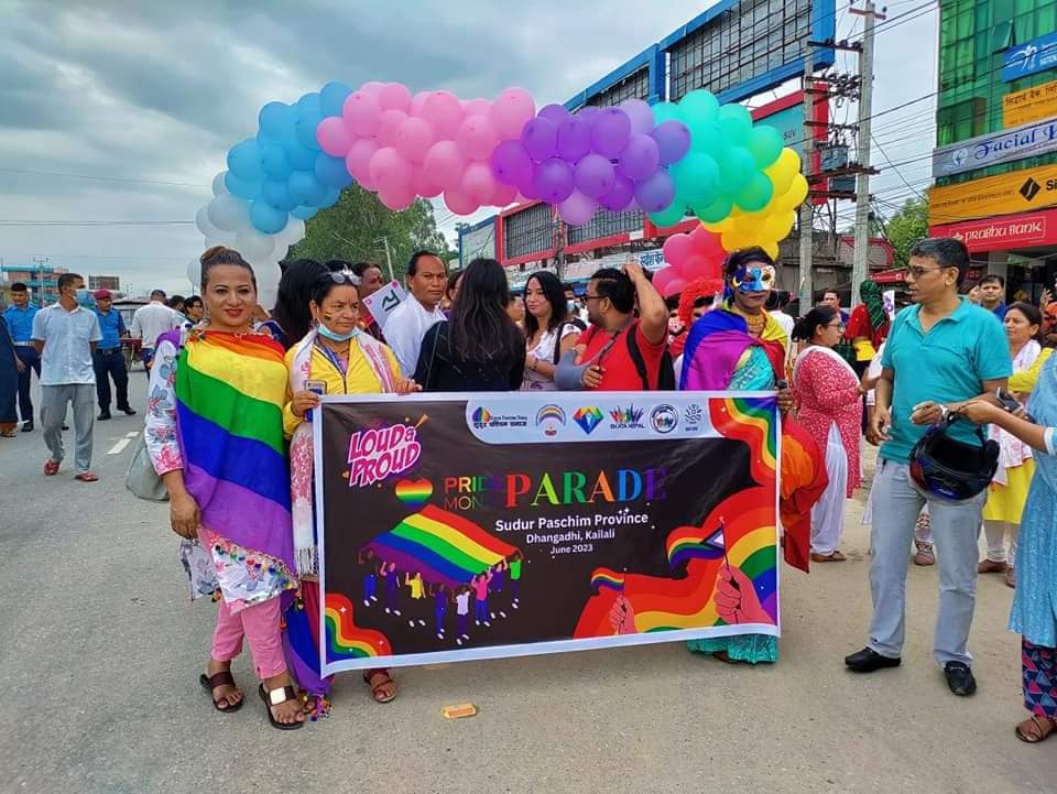 several people in colorful clothing, standing behind a parade banner that says "Pride Month Parade". Behind them is a banner of multi-colored balloons